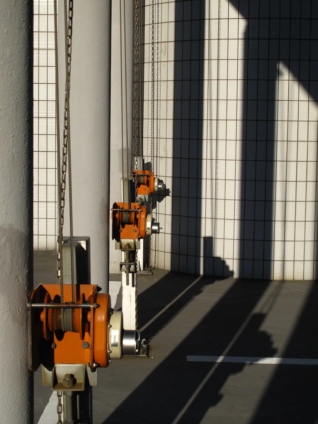 Orange chain hoists lined along a white tiled wall, their shadows cutting diagonally across the floor, Japan