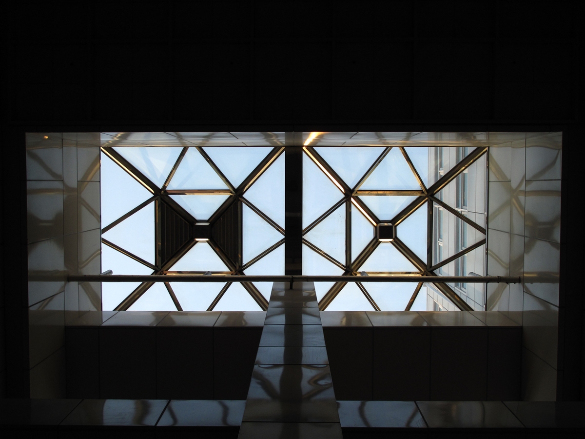 Looking up through two geometric skylight panels in dark steel framing against a bright sky, Japan