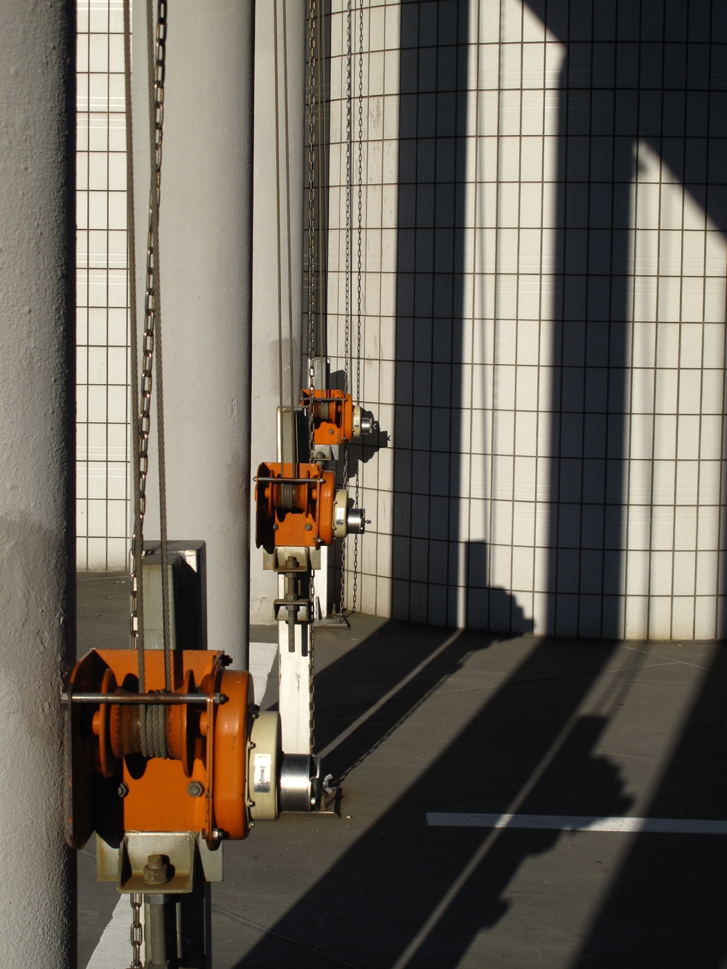 Orange chain hoists lined along a white tiled wall, their shadows cutting diagonally across the floor, Japan